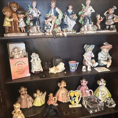Sale Photo Thumbnail #104: Collection of vintage porcelain and ceramic figurines, including Josef Originals Birthday Girls, arranged on dark wooden display shelves.