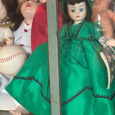 Sale Photo Thumbnail #26: Vintage Madame Alexander-style doll wearing a green dress and matching bonnet, displayed alongside other collectible dolls in a glass cabinet.