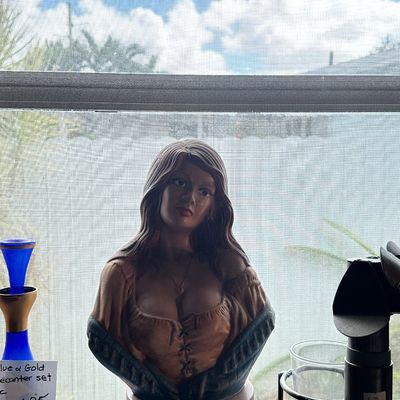 Sale Photo Thumbnail #29: Vintage ceramic bust of a woman with long hair and low-cut bodice, displayed on a wooden pedestal base alongside a cobalt blue glass decanter.