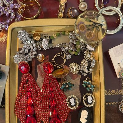 Sale Photo Thumbnail #80: Assortment of vintage costume jewelry, including oversized red rhinestone statement earrings, cameo brooches, clip-on earrings, and gold-tone pieces arranged on a small metal tray.