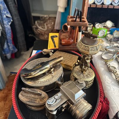 Sale Photo Thumbnail #25: Assortment of vintage novelty table lighters, including a brass Nikon camera lighter and a cherub-adorned lighter, alongside pipes, pocket knives, and watches.