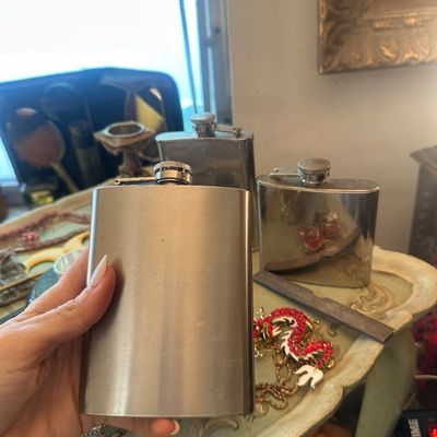 Sale Photo Thumbnail #74: Hand holding an engraved stainless steel hip flask alongside two additional metal flasks on an ornate side table.