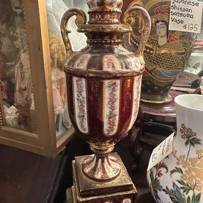 Sale Photo Thumbnail #102: Ornate Victorian-style maroon and gold porcelain urn with a lid and square pedestal base, flanked by Japanese Satsuma and floral porcelain vases.