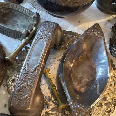 Sale Photo Thumbnail #41: Collection of vintage silver-plated and metal decorative items, featuring an ornate repoussé handset cover, a small marquise dish with floral accents, and a brass pestle resting on a pierced tray.