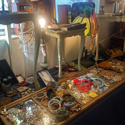 Sale Photo Thumbnail #121: Assortment of vintage costume jewelry, including brooches, necklaces, and bangles, arranged inside a ornate framed mirror display, with small accent tables and a stained glass shade nearby.