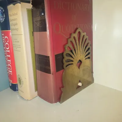 Sale Photo Thumbnail #110: Vintage brass art deco-style bookend holding a collection of reference books, including 'Dictionary of Quotations', on a white shelf.