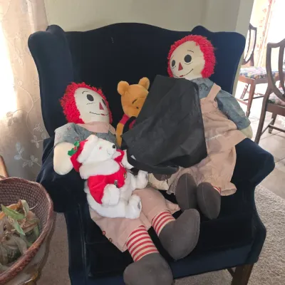 Sale Photo Thumbnail #37: Pair of vintage Raggedy Ann and Andy style cloth dolls seated in a blue velvet wingback armchair alongside Winnie the Pooh and Christmas bear plushies.