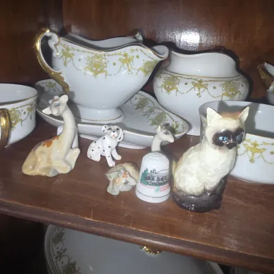Sale Photo Thumbnail #89: Collection of miniature porcelain animal figurines, including a Siamese cat, giraffe, and Dalmatian, displayed alongside gold-accented china on a wooden cabinet shelf.