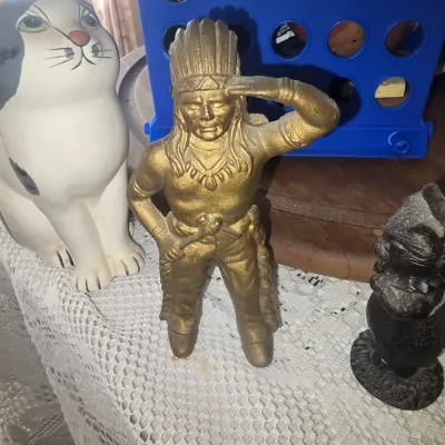 Sale Photo Thumbnail #83: Vintage gold-tone cast metal Native American chief figure alongside a large ceramic cat statue on a lace table runner.