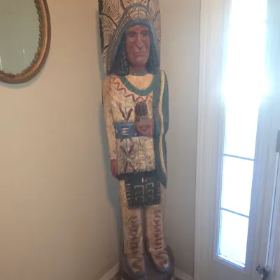 Sale Photo Thumbnail #62: Hand-carved and painted wooden Native American chief statue wearing a feathered headdress, standing in an entryway.