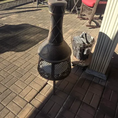 Sale Photo Thumbnail #101: Cast iron chiminea with diamond lattice texture and wire mesh screen, sitting on a brick patio alongside a metal basket filled with firewood.
