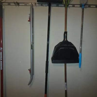 Sale Photo Thumbnail #76: Assorted long-handled lawn, garden, and cleaning tools hanging on a wall track, including a Garden Weasel cultivator, Pittsburgh level, push broom, lawn rake, and dustpan.