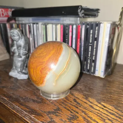 Sale Photo Thumbnail #33: Polished banded agate or onyx sphere displayed on a clear stand, positioned in front of a row of compact discs.
