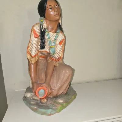 Sale Photo Thumbnail #46: Hand-painted molded ceramic figurine depicting a kneeling Native American woman holding a water jar, resting on a flat white surface.