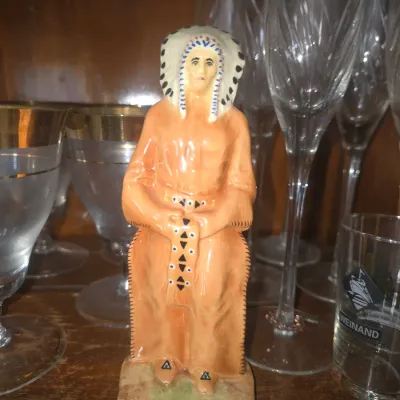 Sale Photo Thumbnail #91: Vintage painted ceramic figurine depicting a Native American chief wearing a feather headdress, displayed in a cabinet alongside glassware.