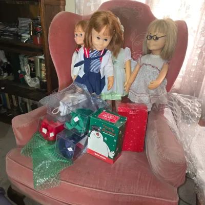 Sale Photo Thumbnail #36: Collection of vintage dolls and boxed holiday collectibles arranged on a pink velvet tufted armchair.