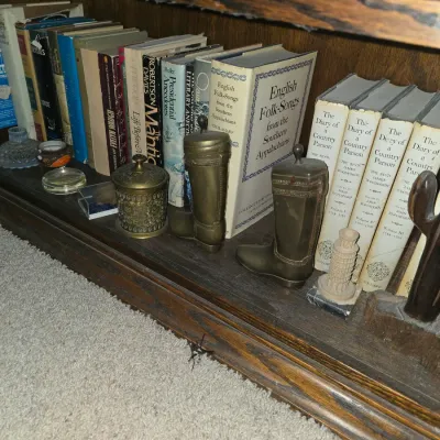 Sale Photo Thumbnail #30: A shelf of vintage books accompanied by brass boot-shaped trinket containers, an ornate lidded pot, and small decorative collectibles.