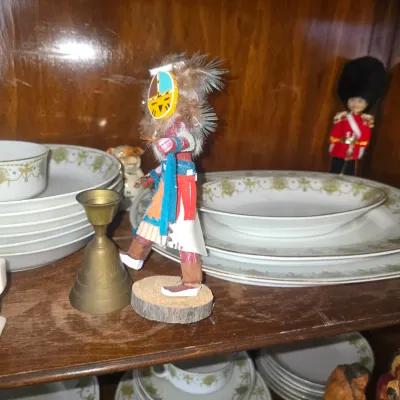 Sale Photo Thumbnail #87: Hand-carved Hopi Katsina doll featuring feather detailing alongside vintage brass candlesticks and fine china inside a wooden hutch.