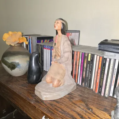 Sale Photo Thumbnail #34: Hand-painted Native American style kneeling woman figurine alongside a dark stone seal sculpture and a glazed studio pottery vase, set on a wooden shelf lined with CDs.