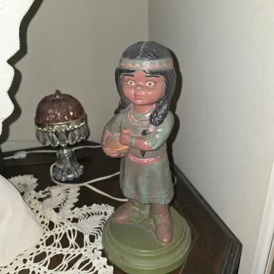 Sale Photo Thumbnail #49: Vintage painted chalkware or plaster figurine of a Native American girl holding a pot, displayed on a wooden table beside a small glass boudoir lamp.