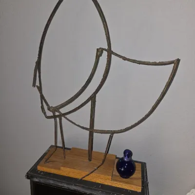 Sale Photo Thumbnail #25: Abstract mid-century style wire sculpture of a bird mounted on a wooden base, displayed alongside a cobalt blue glass bottle on a small table.