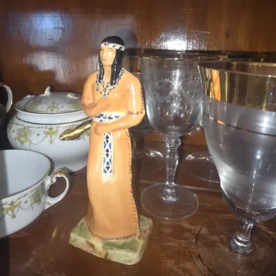 Sale Photo Thumbnail #90: Hand-painted ceramic figurine of a Native American woman mounted on a marble base, displayed alongside etched and gold-rimmed glassware inside a wooden hutch.