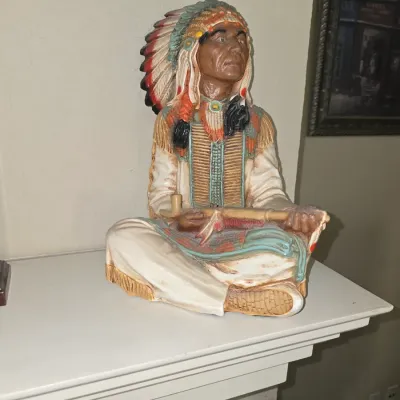 Sale Photo Thumbnail #45: Hand-painted composite or resin figurine of a seated Native American chief holding a ceremonial pipe, detailed with a full feather headdress and traditional attire.
