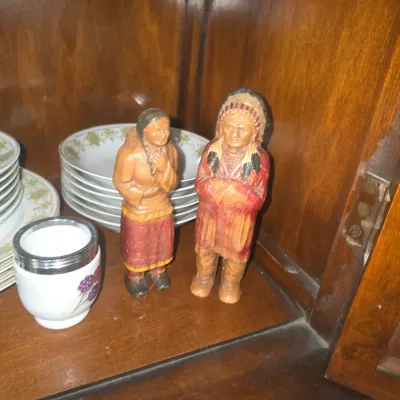Sale Photo Thumbnail #86: Pair of vintage carved and painted Native American figurines displayed in a wooden cabinet alongside assorted china plates and a small cup.
