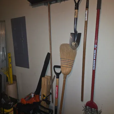 Sale Photo Thumbnail #75: Collection of long-handled yard and garden tools mounted on a garage wall, including a Garden Weasel cultivator, HDX shovel, corn broom, and push broom.