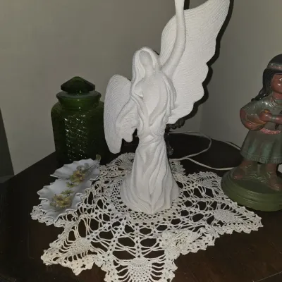 Sale Photo Thumbnail #50: White textured ceramic angel figurine with raised wing displayed on a crocheted lace doily, alongside a green glass jar and a small floral dish.