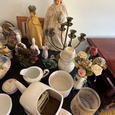 Sale Photo Thumbnail #55: A collection of vintage home decor items including religious statues of Infant Jesus of Prague, a brass candelabra, porcelain teaware, and artificial flower arrangements.
