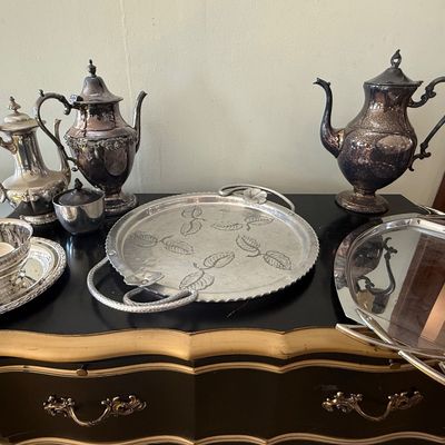 Sale Photo Thumbnail #42: Collection of vintage silver-plated serving pieces, including teapots, coffee pots, trays, and a small covered sugar bowl displayed on a black buffet table.