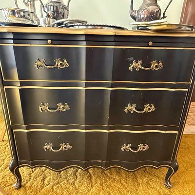 Sale Photo Thumbnail #43: French Provincial style black painted three-drawer dresser with gold trim accents and ornate brass hardware, topped with assorted silver-plated serving pieces.