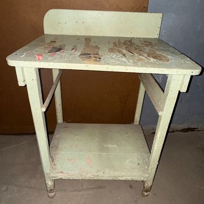 Sale Photo Thumbnail #11: Vintage pale green painted wooden utility stand with a back backsplash and lower shelf, displaying heavy surface wear, peeling paint, and newspaper residue on the tabletop.