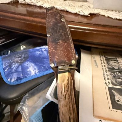 Vintage hunting knife featuring a stag horn handle and a brass handguard, stored inside a worn brown leather sheath with metal rivets.
