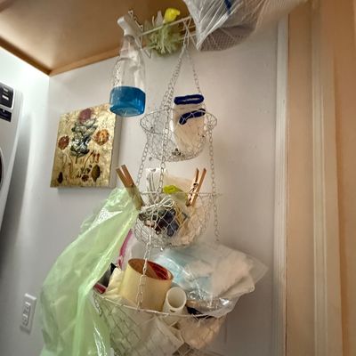 Three-tier white wire hanging fruit or storage basket filled with household cleaning supplies and odds and ends.