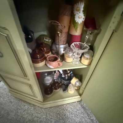 Collection of decorative candles, glass holders, and pottery pieces stored inside a painted sage green corner cabinet.