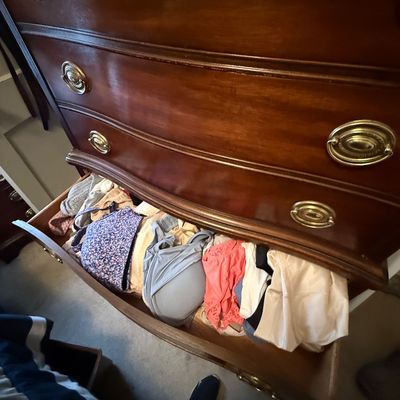 Traditional dark wood tall chest of drawers with brass oval pull handles, showing an open drawer filled with women's undergarments.