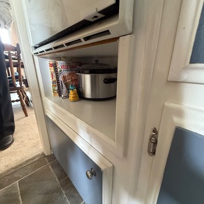 Oval stainless steel slow cooker with a glass lid sitting inside a kitchen cabinet cubby alongside cookbooks.