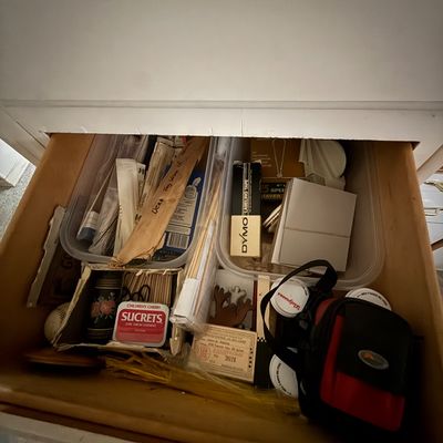Open wooden drawer filled with household junk drawer items, including a vintage Sucrets tin, a box of Dymo labeling tape, plastic storage...