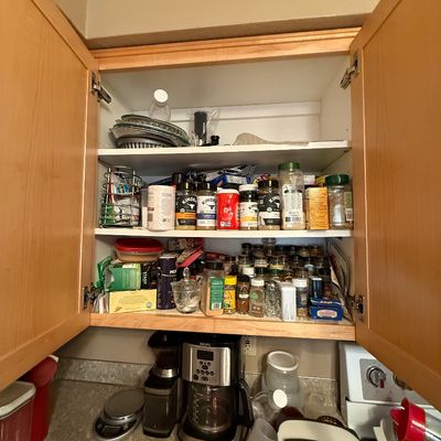 Open kitchen cabinet stocked with various spices, seasonings, pie tins, and food containers above a countertop with a Krups coffee maker.