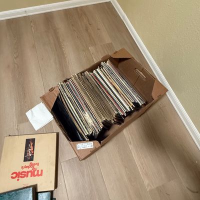 Cardboard box filled with vinyl LP records sitting on a wood-look floor, accompanied by vintage record albums nearby.