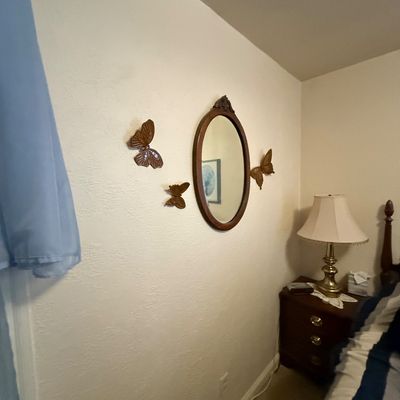 Vintage oval wood-framed wall mirror surrounded by a three-piece set of carved wood butterfly wall plaques, displayed above a bedside table.