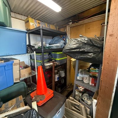 Crowded shed containing plastic shelving units filled with various storage totes, paint cans, tool supplies, and an orange safety cone.