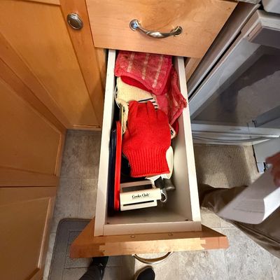 Open kitchen cabinet drawer filled with various kitchen linens and accessories, including a red oven mitt, potholders, and a Cooks Club...