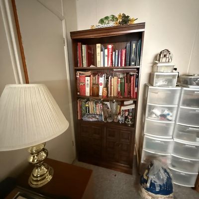 Tall dark wood bookcase filled with assorted hardcovers, paperbacks, and decorative knick-knacks, positioned next to a brass table lamp...