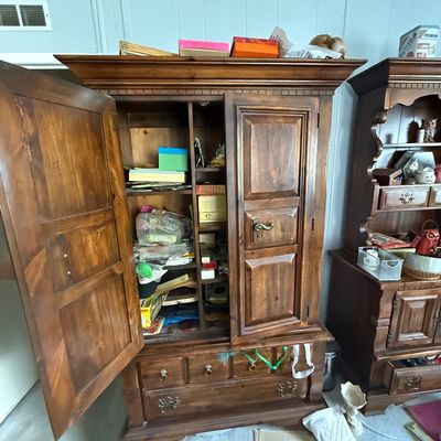 Large rustic pine armoire with panel doors, lower drawers, and brass hardware, filled with various household odds and ends.