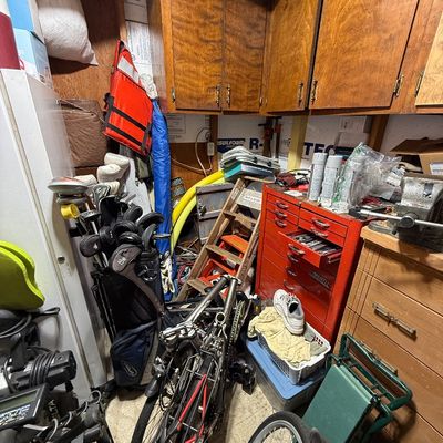 Cluttered workshop and storage area featuring a red Cornwell tool chest, bicycles, golf clubs with bags, a wooden stepladder, and...