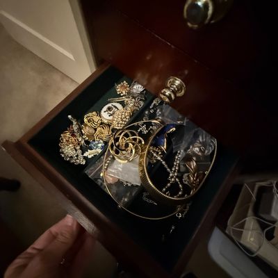 Open wooden jewelry armoire drawer filled with an assortment of vintage costume jewelry, including gold-tone brooches, beaded necklaces,...