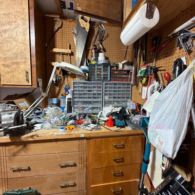 Workshop corner with wooden drawers, pegboard storage, hand saws, hardware organizers, and an articulated desk lamp.